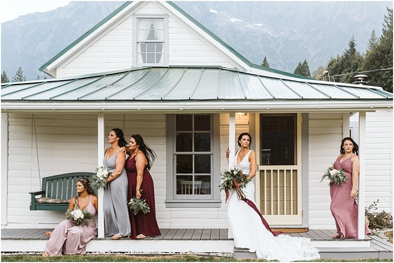 Premier snohomish & seattle wedding photographer | gsquared weddings photography five women in formal dresses including a bride in white holding a bouquet pose on the porch of a white house with a green roof near snohomish set against a scenic mountain and tree backdrop | serving snohomish seattle                   & beyond