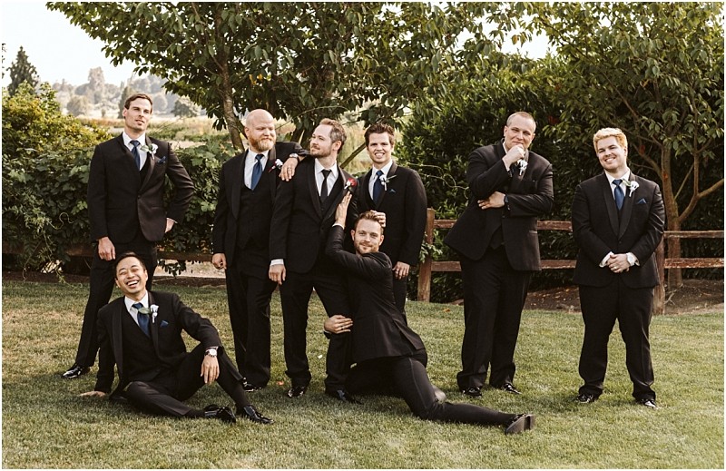 Premier snohomish & seattle wedding photographer | gsquared weddings photography a group of eight men in black suits and ties pose playfully outdoors on grass at a snohomish seattle wedding with trees and a wooden fence in the background Some stand smiling while others strike humorous relaxed poses in front | serving snohomish seattle                   & beyond