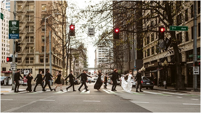 Premier Snohomish & Seattle Wedding Photographer | GSquared Weddings Photography A wedding party crosses a city street at a crosswalk, hand in hand, with the bride and groom at the center, surrounded by attendants in formal black attire. Tall buildings line the scene during this unforgettable Seattle wedding.                                   | Serving Snohomish, Seattle ,                  & Beyond