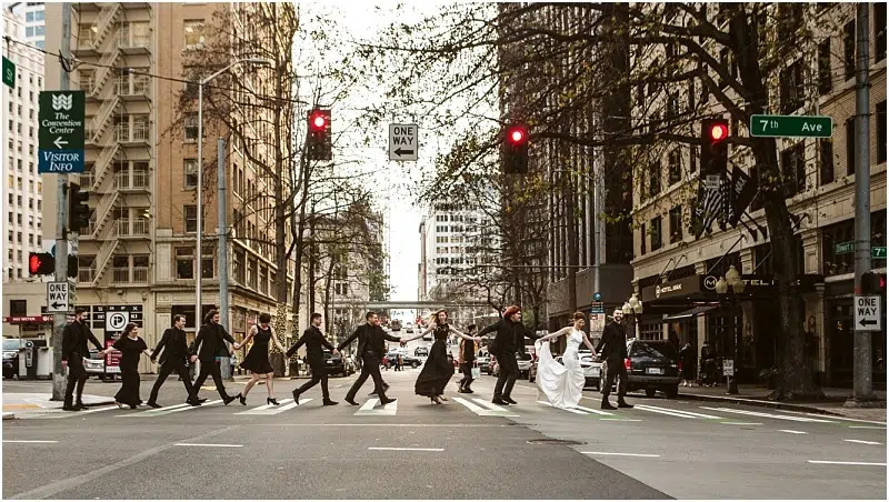Premier Snohomish & Seattle Wedding Photographer | GSquared Weddings Photography A wedding party crosses a city street at a crosswalk, hand in hand, with the bride and groom at the center, surrounded by attendants in formal black attire. Tall buildings line the scene during this unforgettable Seattle wedding. 47.9129° N, 122.0982° W | Serving Snohomish, Seattle , Orlando & Beyond