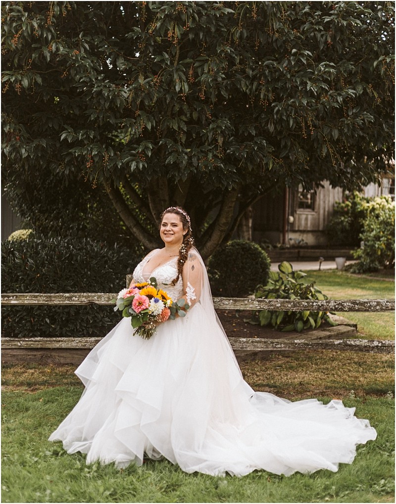 Premier snohomish & seattle wedding photographer | gsquared weddings photography a bride in a white layered wedding gown stands on grass near snohomish smiling and holding a vibrant bouquet She has long braided hair and is posed in front of a large leafy tree with rustic buildings in the background | serving snohomish seattle                   & beyond