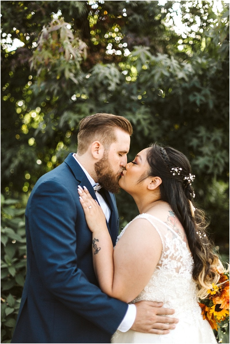 Premier snohomish & seattle wedding photographer | gsquared weddings photography a snohomish bride and groom share a kiss outdoors surrounded by greenery The groom wears a navy suit and the bride has a lace dress with flowers in her hair holding a bouquet Sunlight filters through the leaves behind them | serving snohomish seattle                   & beyond