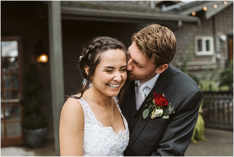 Premier snohomish & seattle wedding photographer | gsquared weddings photography a groom in a suit kisses a smiling bride on the cheek during their joyful snohomish outdoor wedding both appearing affectionate The bride wears a white dress and the groom highlights his look with a red rose boutonniere | serving snohomish seattle                   & beyond