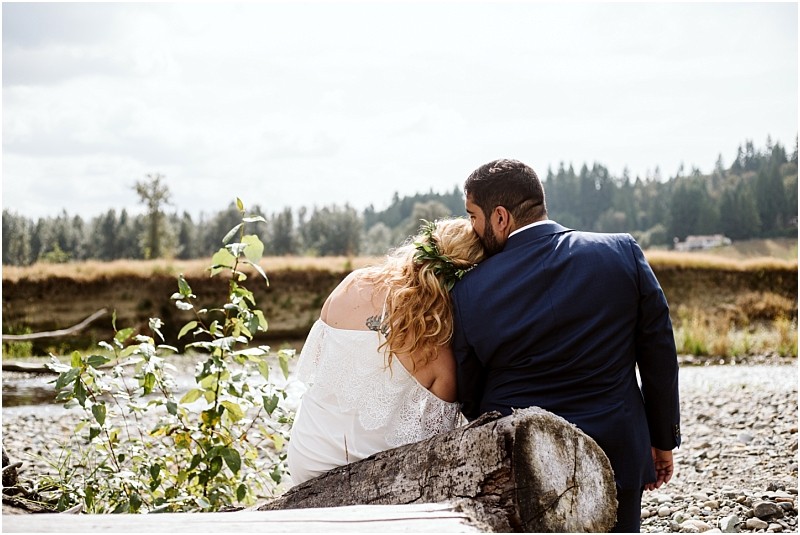 Snohomish & seattle wedding photographer | gsquared weddings photography a couple featured in best of 2019 wedding couples sits on a fallen log by a rocky riverbank the bride resting her head on the grooms shoulder as they gaze at a scenic tree lined landscape | wedding photo by gsquared weddings