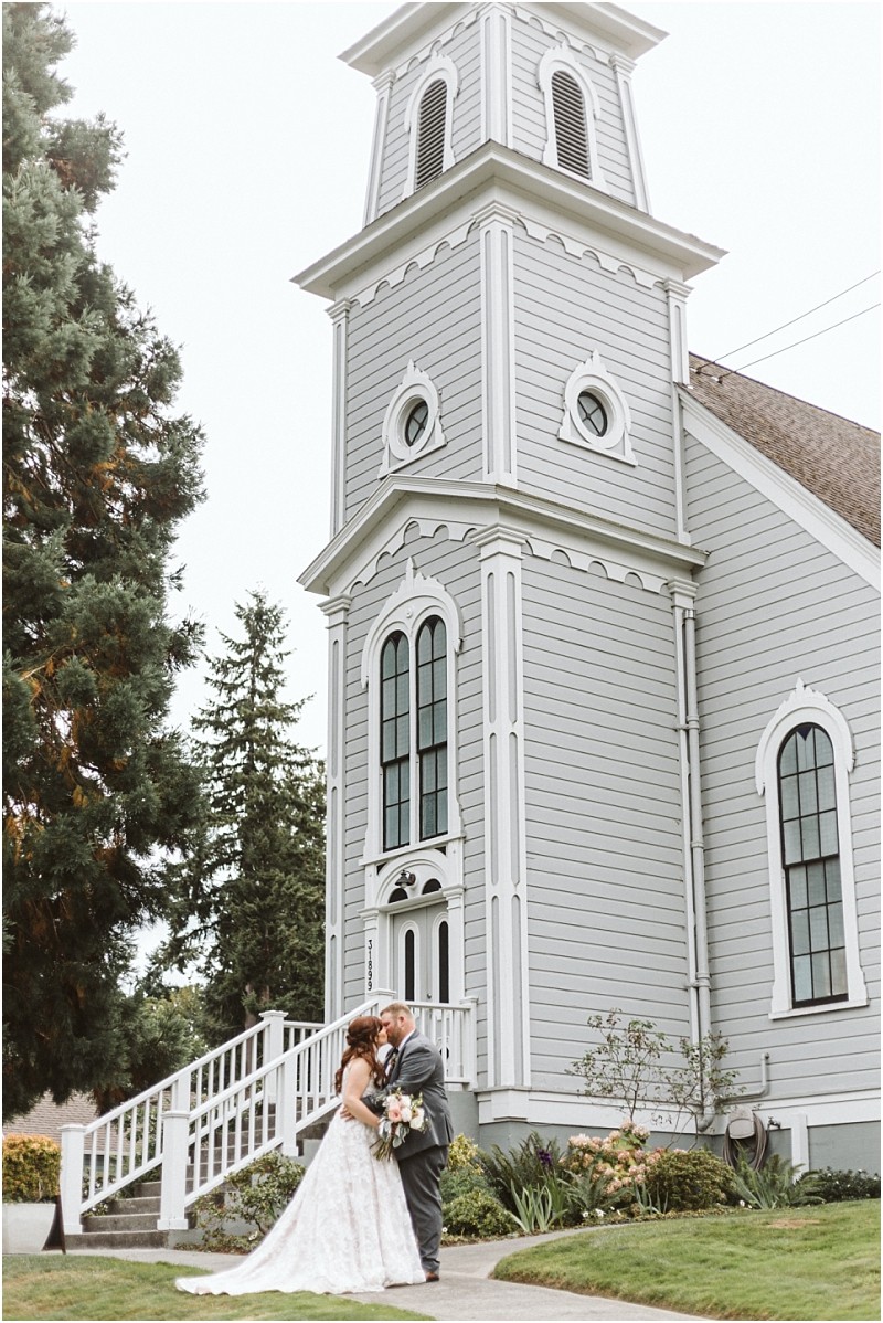 Snohomish & seattle wedding photographer | gsquared weddings photography wedding couples share an intimate moment outside a tall gray wooden church with arched windows standing together on a sidewalk surrounded by greenerycapturing the best of 2019 romance | wedding photo by gsquared weddings