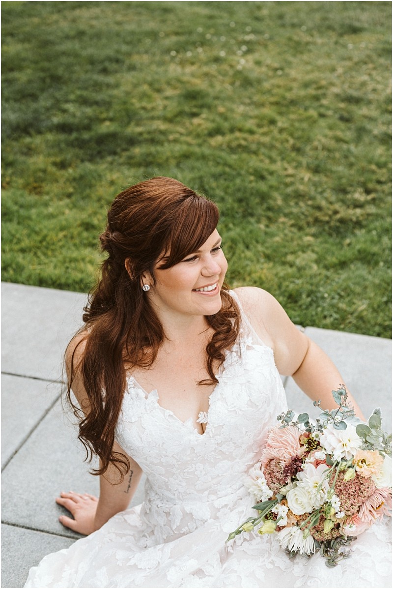 Snohomish & seattle wedding photographer | gsquared weddings photography a bride with long brown hair in loose waves wearing a white lace wedding dress sits on a stone path holding a bouquet of flowers She is smiling and looking off to the side part of our best of 2019 wedding couples collection | wedding photo by gsquared weddings