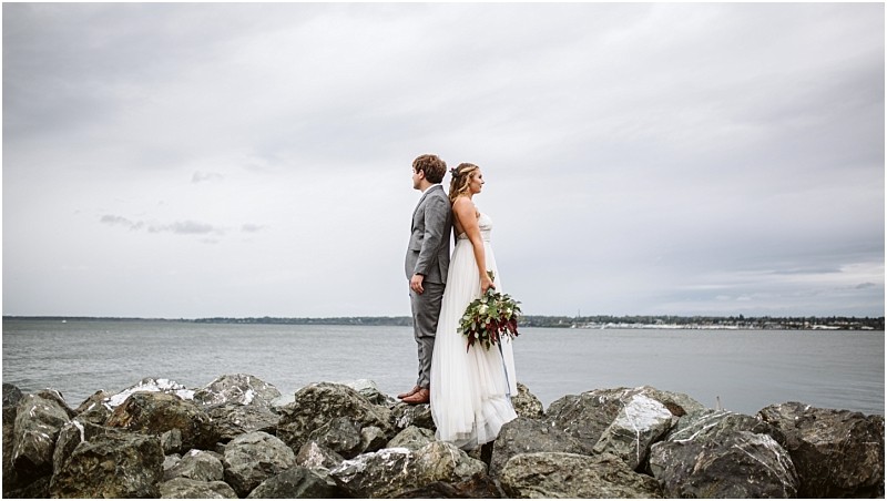 Snohomish & seattle wedding photographer | gsquared weddings photography a bride and groom stand back to back on a rocky shore facing away from each other with the ocean and cloudy sky in the background This best of 2019 moment captures wedding couples in a stunning seaside setting | wedding photo by gsquared weddings