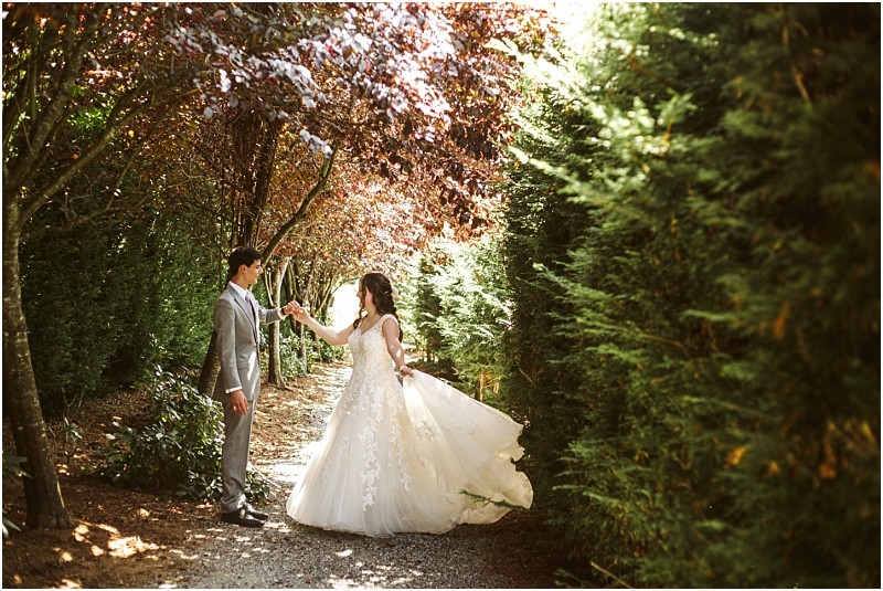 Snohomish & seattle wedding photographer | gsquared weddings photography a bride in a white gown and a groom in a gray suit hold hands on a sunlit garden path surrounded by lush green trees and dappled sunlighta true best of 2019 moment for wedding couples | wedding photo by gsquared weddings