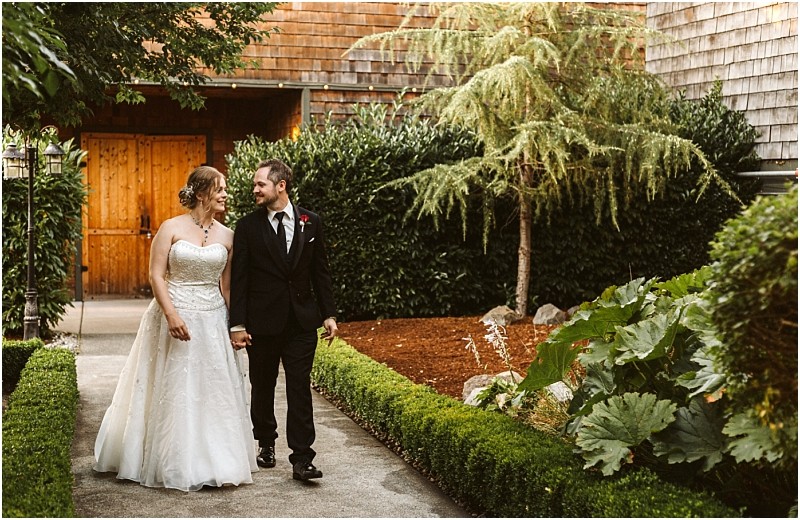 Snohomish & seattle wedding photographer | gsquared weddings photography one of our best of 2019 wedding couples a bride in a white dress and a groom in a black suit walk hand in hand on a garden path smiling at each other with lush greenery and a charming wooden building behind them | wedding photo by gsquared weddings