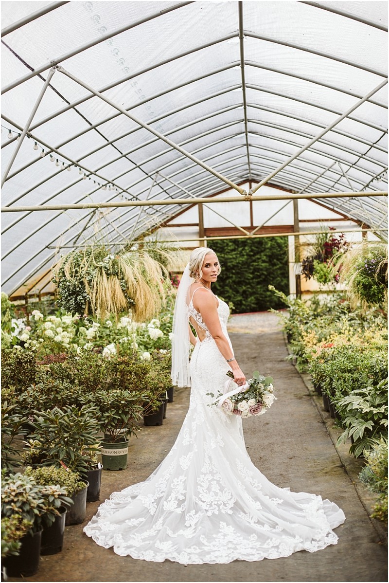 Snohomish & seattle wedding photographer | gsquared weddings photography a bride in a white lace gown and veil stands holding a bouquet in a greenhouse filled with lush green plants and flowers looking over her shoulder at the cameracapturing the magic of wedding couples best of 2019 | wedding photo by gsquared weddings