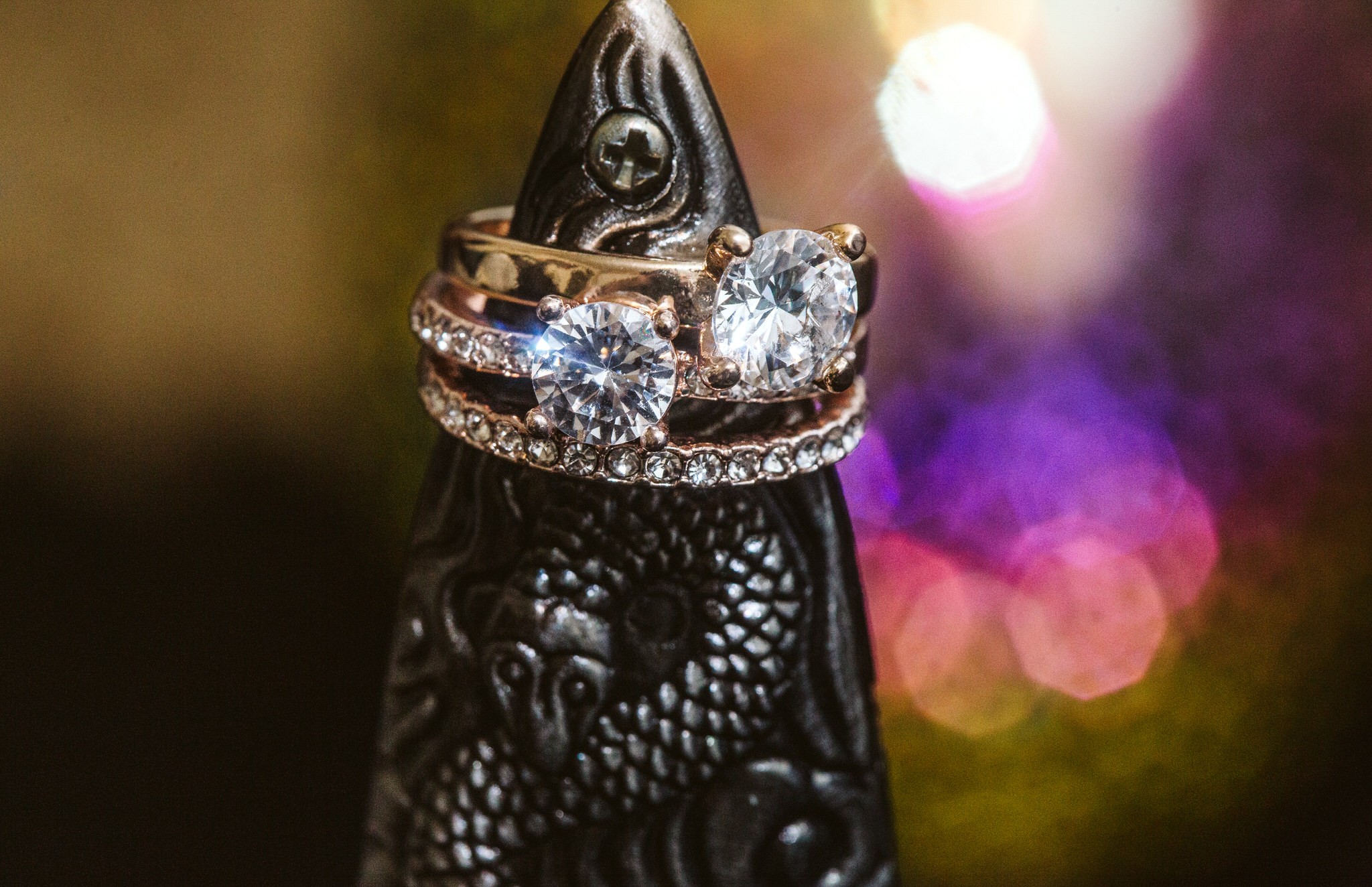 Snohomish & Seattle Wedding Photographer | GSquared Weddings Photography Two diamond rings—one with a row of small diamonds, another with two large stones—rest on a dark, intricately carved object featuring a dragon motif. Blurred colorful lights glow in the background. See more in The Weducation Series: 15 Things You Shouldn't Do on Your Wedding Day.                                   | wedding photo by GSquared Weddings