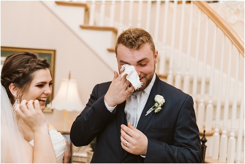 Snohomish & seattle wedding photographer | gsquared weddings photography a groom in a suit wipes away tears with a tissue while smiling standing next to a laughing bride in her wedding dress at their joyful marysville wedding both captured in front of a staircase | wedding photo by gsquared weddings