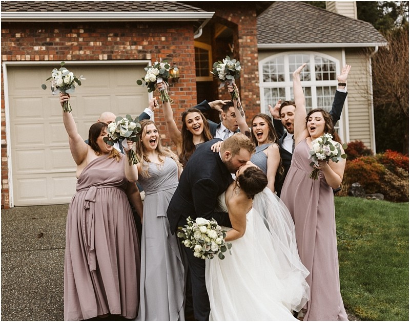 Snohomish & seattle wedding photographer | gsquared weddings photography a bride and groom kiss in front of a house during their marysville wedding while their wedding party dressed in lavender and gray dresses cheers and raises bouquets celebrating joyfully on a cloudy day | wedding photo by gsquared weddings
