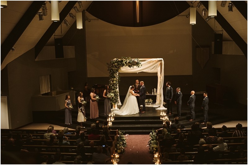 Snohomish & seattle wedding photographer | gsquared weddings photography a bride and groom stand under a floral arch exchanging vows on a decorated stage surrounded by their wedding party in a dimly lit church as guests watch from the pews at their beautiful marysville wedding | wedding photo by gsquared weddings
