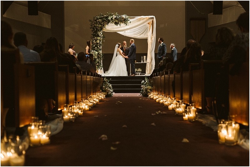 Snohomish & seattle wedding photographer | gsquared weddings photography a bride and groom stand at the altar under a floral arch in a dimly lit marysville wedding venue Guests are seated on either side of an aisle lined with glowing candles in glass jars and rose petals | wedding photo by gsquared weddings