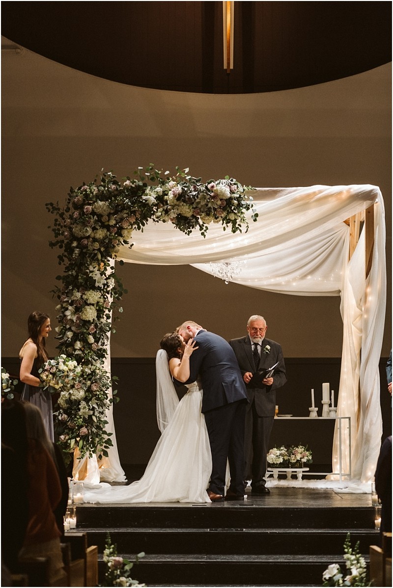Snohomish & seattle wedding photographer | gsquared weddings photography a bride and groom share a kiss beneath a floral wedding arch at their marysville wedding while an officiant stands nearby and guests watch The ceremony setting is beautifully decorated with flowers and draped white fabric | wedding photo by gsquared weddings
