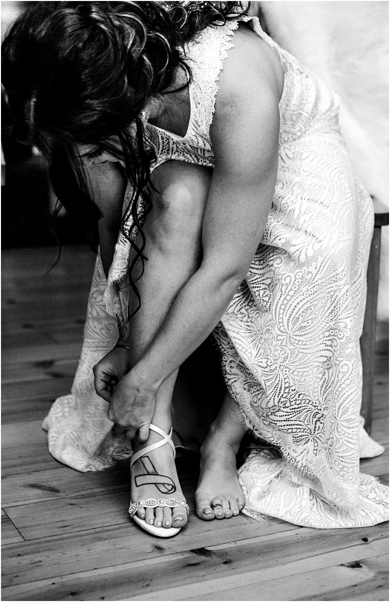Snohomish & seattle wedding photographer | gsquared weddings photography a woman in a lace wedding dress sits on a chair bending forward to fasten the strap of her white heeled sandal One foot is bare on the wooden floor This elegant black and white image captures a quiet moment before her leavenworth wedding | wedding photo by gsquared weddings