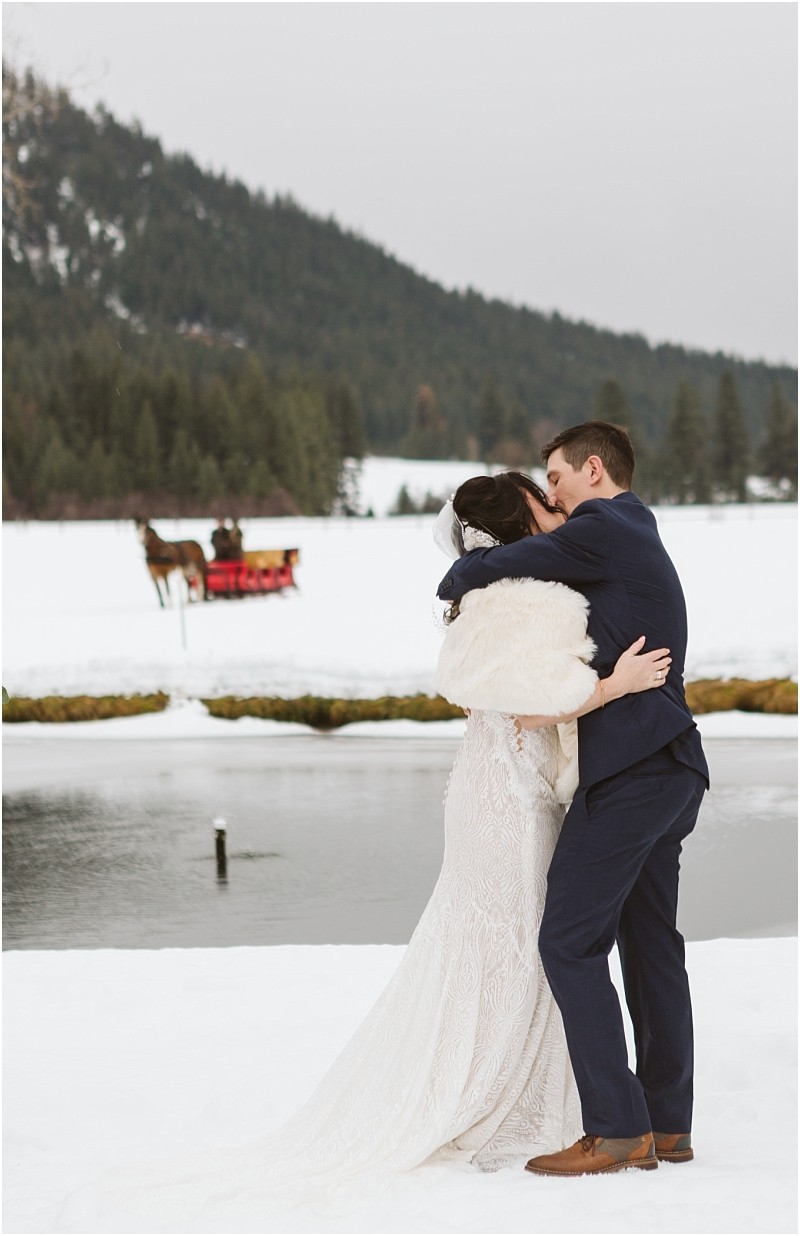 Snohomish & seattle wedding photographer | gsquared weddings photography a bride and groom embrace and kiss in the snow by a pond with a wintry forest and mountains in the backgrounda magical leavenworth wedding complete with a horse drawn sleigh carrying passengers in the distance | wedding photo by gsquared weddings
