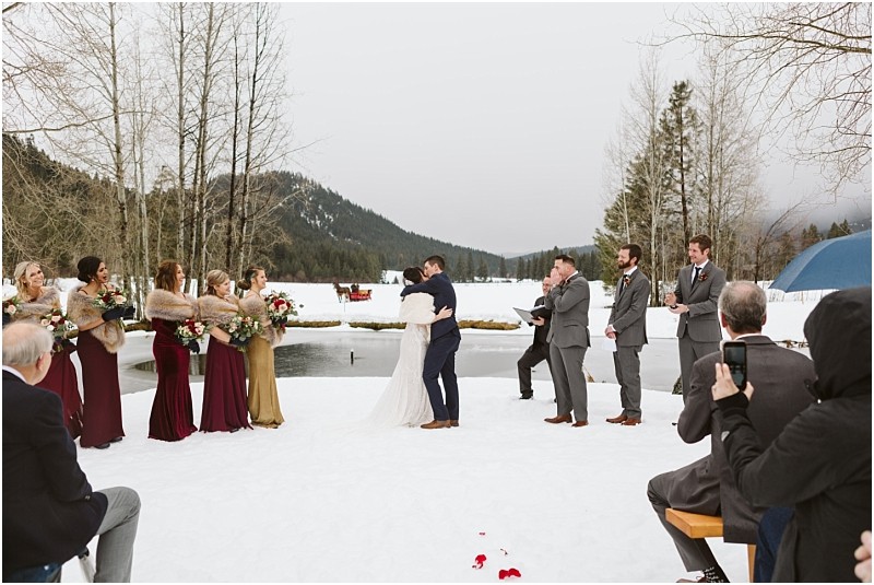 Snohomish & seattle wedding photographer | gsquared weddings photography a bride and groom kiss at their outdoor winter leavenworth wedding ceremony Bridesmaids in colorful dresses and groomsmen in gray suits stand on either side as snow covers the ground with guests surrounded by trees and mountains | wedding photo by gsquared weddings