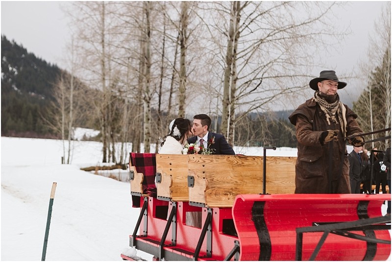 Snohomish & seattle wedding photographer | gsquared weddings photography a couple in formal attire shares a kiss during their leavenworth wedding riding in a wooden sleigh pulled by a man in winter clothes and a cowboy hat through a snowy forested landscape | wedding photo by gsquared weddings