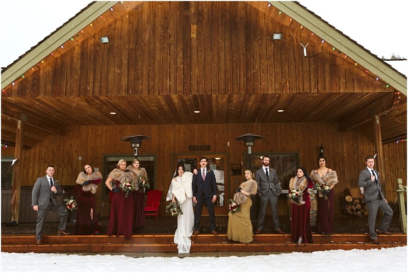 Snohomish & seattle wedding photographer | gsquared weddings photography a wedding party poses on the porch of a wooden cabin in winter during a leavenworth wedding The bride and groom are at the center bridesmaids in burgundy dresses and groomsmen in gray suits stand on either side with snow covering the ground | wedding photo by gsquared weddings