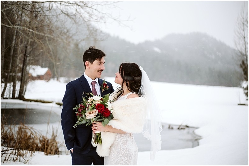 Snohomish & seattle wedding photographer | gsquared weddings photography a bride and groom stand outdoors in the snow at their leavenworth wedding smiling at each other The bride holds a red and white bouquet and wears a veil and fur shawl while snowy trees and a pond complete the wintry scene | wedding photo by gsquared weddings