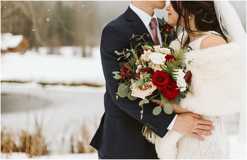 Snohomish & seattle wedding photographer | gsquared weddings photography a bride and groom embrace outdoors in winter during their leavenworth wedding The bride holds a bouquet of red and white flowers and wears a white fur stole while the groom is in a dark suit amid snow covered trees | wedding photo by gsquared weddings