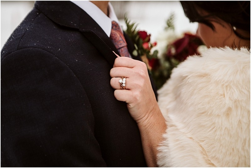 Snohomish & seattle wedding photographer | gsquared weddings photography a close up of a couple embracing at a leavenworth wedding focusing on the womans hand with a diamond ring resting on a mans suit lapel She wears a fur coat and holds a bouquet of red flowers in the background | wedding photo by gsquared weddings