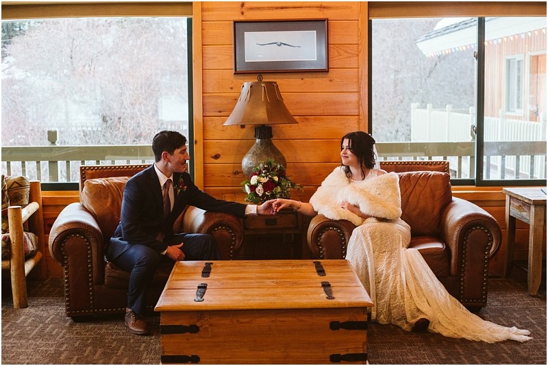 Snohomish & seattle wedding photographer | gsquared weddings photography a bride in a white dress and fur shawl sits on a leather armchair holding hands with her groom in a suit on another armchair inside a cozy wooden cabin with large windowscapturing the charm of an intimate leavenworth wedding | wedding photo by gsquared weddings