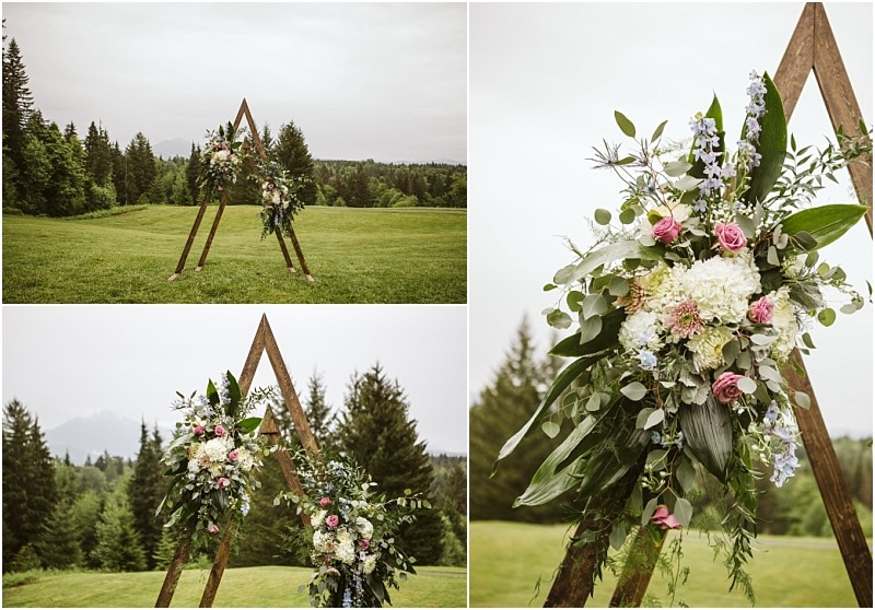 Snohomish & seattle wedding photographer | gsquared weddings photography a triangular wooden wedding arch perfect for a machias elopement is adorned with white and pink flowers and greenery in a grassy field Surrounded by trees under a cloudy sky close ups reveal the floral detailspure love with a view | wedding photo by gsquared weddings