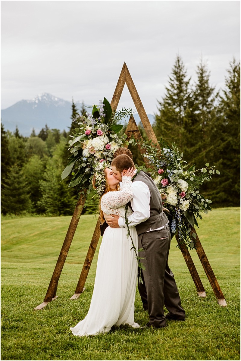 Snohomish & seattle wedding photographer | gsquared weddings photography a bride and groom share a kiss in front of a wooden triangular wedding arch adorned with flowers and greenery set outdoors on a grassy lawnperfect for a machias elopement celebrating love with a view of trees and mountains | wedding photo by gsquared weddings