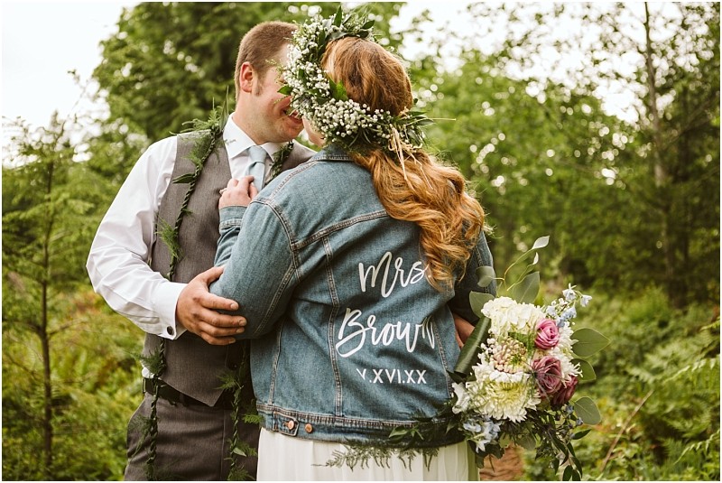 Snohomish & seattle wedding photographer | gsquared weddings photography a bride and groom embrace outdoors at their machias elopement The bride in a denim jacket reading mrs Brown and a floral crown holds her bouquet as they share an intimate moment surrounded by lush greenerylove with a view | wedding photo by gsquared weddings