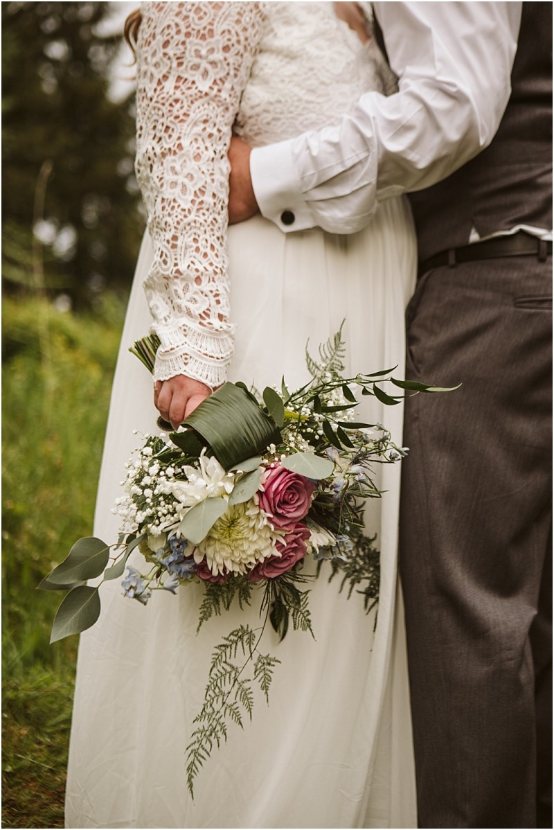 Snohomish & seattle wedding photographer | gsquared weddings photography a bride in a lace wedding dress holds a bouquet of purple white and blue flowers while standing close to her groom in a white shirt and gray vestcelebrating love with a view at their machias elopement outdoors | wedding photo by gsquared weddings