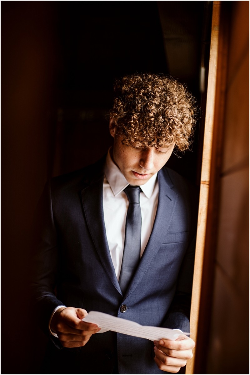 Snohomish & seattle wedding photographer | gsquared weddings photography a young man with curly hair dressed in a dark suit and tie stands indoors by a wooden wall at marionfield farm elopement looking down thoughtfully at a piece of paper he is holding in both hands | wedding photo by gsquared weddings