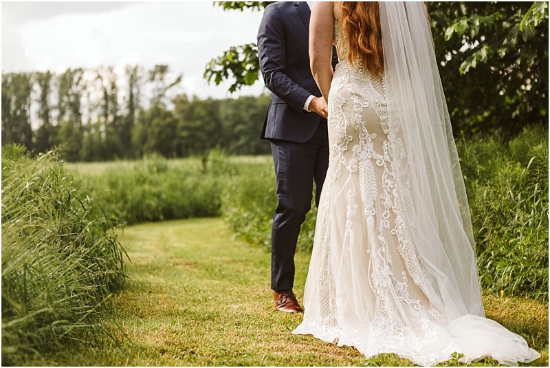 Snohomish & seattle wedding photographer | gsquared weddings photography a bride in a lace wedding gown and veil holds hands with a groom in a navy suit as they stand on a grassy path at their marionfield farm elopement surrounded by greenery and trees on a sunny day | wedding photo by gsquared weddings
