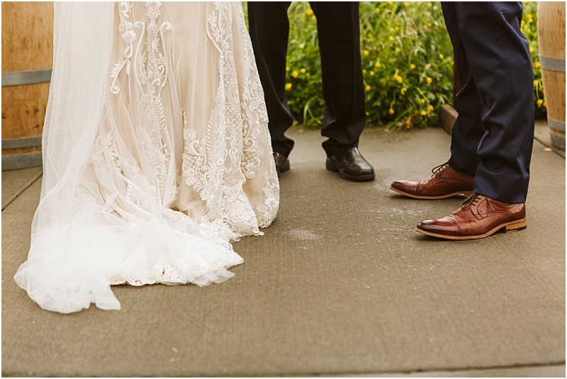 Snohomish & seattle wedding photographer | gsquared weddings photography close up of a brides lace wedding dress and two mens dress shoesone black one brownstanding on concrete outdoors with greenery and yellow flowers in the background at a marionfield farm elopement | wedding photo by gsquared weddings