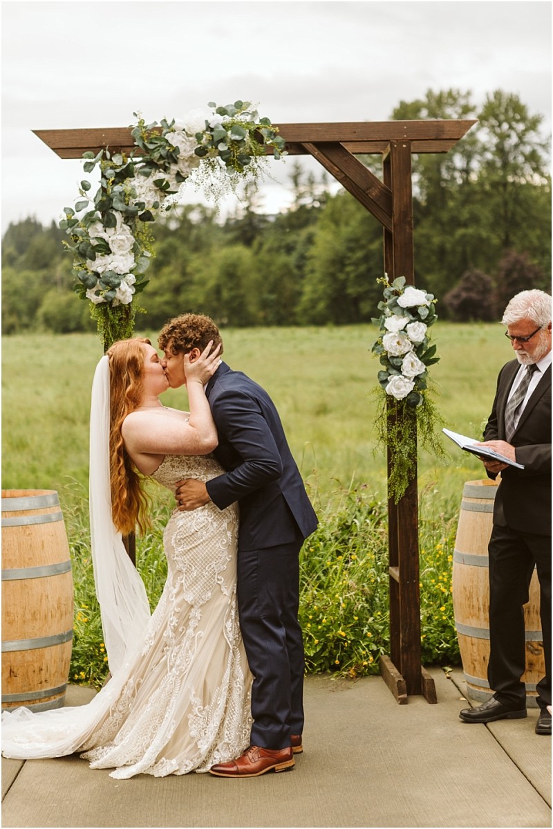 Snohomish & seattle wedding photographer | gsquared weddings photography a bride and groom share a kiss under a wooden wedding arch decorated with white flowers and greenery at their marionfield farm elopement outdoors with green fields in the background An officiant stands nearby holding a book | wedding photo by gsquared weddings