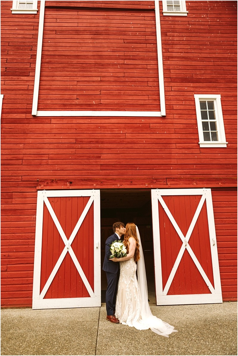 Snohomish & seattle wedding photographer | gsquared weddings photography a bride and groom stand closely together in front of large red barn doors at marionfield farm elopement with the bride holding a bouquet and the groom in a blue suit The barn has white trim and a rustic charming appearance | wedding photo by gsquared weddings