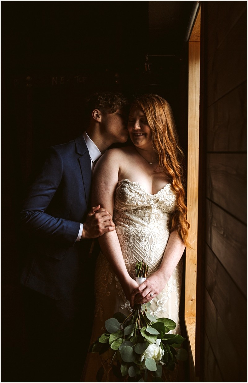 Snohomish & seattle wedding photographer | gsquared weddings photography a groom in a blue suit kisses the brides temple as she smiles holding a bouquet by a wooden window at their marionfield farm elopement The bride wears a strapless lace dress with long wavy red hair bathed in warm intimate lighting | wedding photo by gsquared weddings