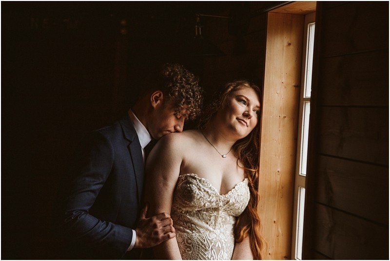 Snohomish & seattle wedding photographer | gsquared weddings photography a bride in a lace dress stands by a window at marionfield farm elopement gazing out with a soft expression while her groom in a suit gently rests his head on her shoulder in the warmly lit room | wedding photo by gsquared weddings
