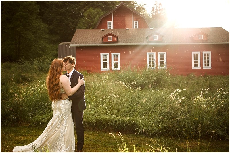 Snohomish & seattle wedding photographer | gsquared weddings photography a bride and groom embrace in tall grass in front of a red barn at their marionfield farm elopement with sunlight streaming through the trees and gentle rain falling creating a romantic and dreamy atmosphere | wedding photo by gsquared weddings