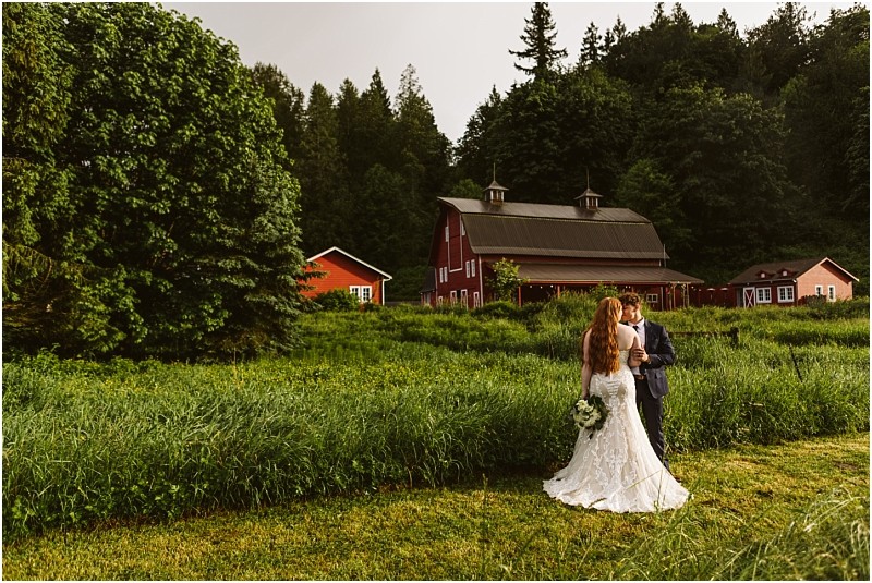 Snohomish & seattle wedding photographer | gsquared weddings photography a bride and groom stand together in a grassy field at marionfield farm facing a rustic red barn and surrounding trees under a partly cloudy sky The bride holds a bouquet and wears a white gown the groom wears a dark suitperfect for an elopement | wedding photo by gsquared weddings