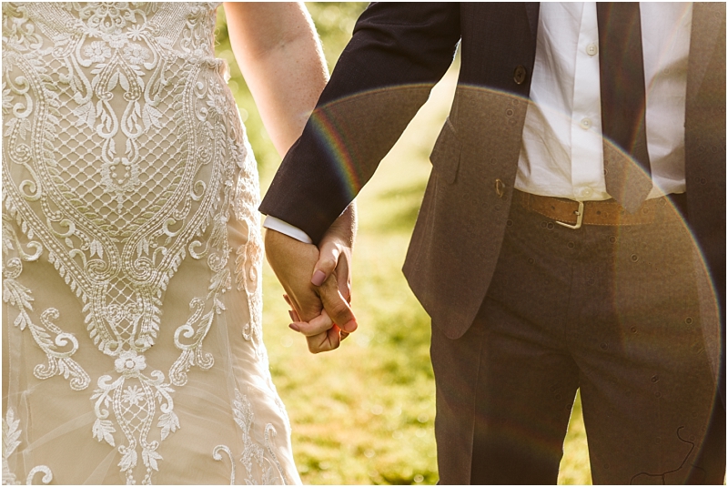 Snohomish & seattle wedding photographer | gsquared weddings photography a close up of a couple holding hands outdoors at their marionfield farm elopement featuring the intricate lace design of a wedding dress and a dark suit sunlight creates a rainbow lens flare across the image | wedding photo by gsquared weddings