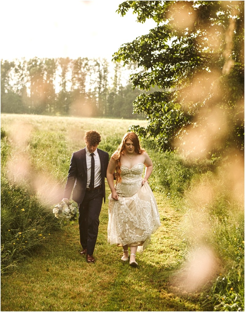 Snohomish & seattle wedding photographer | gsquared weddings photography a couple walks through a sunlit grassy field at marionfield farm elopement The woman in a white lace dress holds up her skirt while the man in a dark suit carries a bouquet Soft sunlight and blurred foliage create a dreamy romantic atmosphere | wedding photo by gsquared weddings
