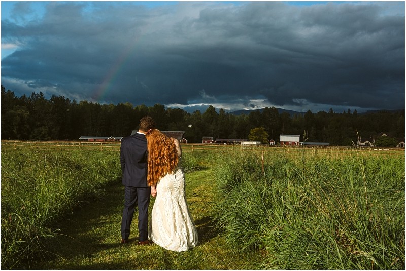 Snohomish & seattle wedding photographer | gsquared weddings photography a bride and groom stand close together in a grassy field at marionfield farm elopement facing away from the camera toward distant barns and trees under a dramatic cloudy sky with a faint rainbow | wedding photo by gsquared weddings
