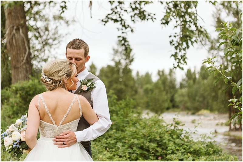 Snohomish & seattle wedding photographer | gsquared weddings photography a groom embraces and kisses the brides head as they stand outdoors in front of green trees and foliage during their methow elopement The bride holds a bouquet and wears a sleeveless gown with a beaded back | wedding photo by gsquared weddings