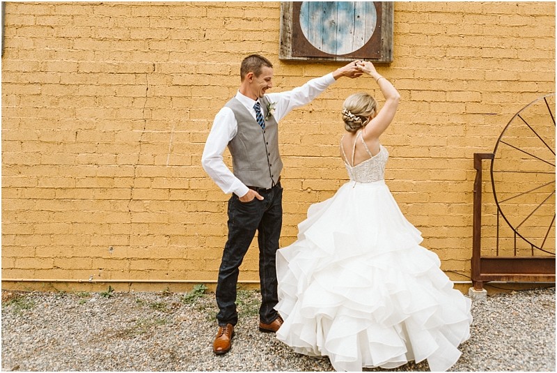 Snohomish & seattle wedding photographer | gsquared weddings photography a groom in a vest and tie twirls his bride who is wearing a white ruffled gown in front of a yellow brick wall decorated with rustic elements during their intimate methow elopement | wedding photo by gsquared weddings