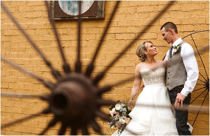 Snohomish & seattle wedding photographer | gsquared weddings photography a bride and groom stand close together smiling in front of a yellow brick wall The image taken during their methow elopement is partially framed by the spokes of a large wagon wheel in the foreground | wedding photo by gsquared weddings