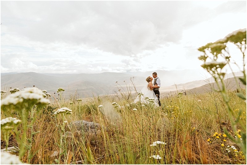 Snohomish & seattle wedding photographer | gsquared weddings photography a couple stands embracing in a field of wildflowers with tall grass mountains and a cloudy sky in the background capturing the romantic and serene spirit of a methow elopement | wedding photo by gsquared weddings