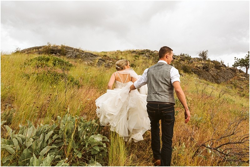 Snohomish & seattle wedding photographer | gsquared weddings photography a bride in a white dress and a groom in a vest walk up a grassy hillside together under a cloudy sky surrounded by wild plants and rocky terraina beautiful methow elopement | wedding photo by gsquared weddings
